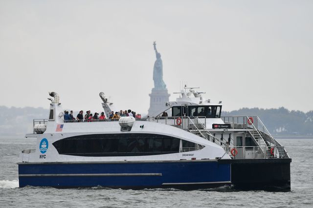 A NYC Ferry cruises on the Hudson River, passing the Statue of Liberty in the background.